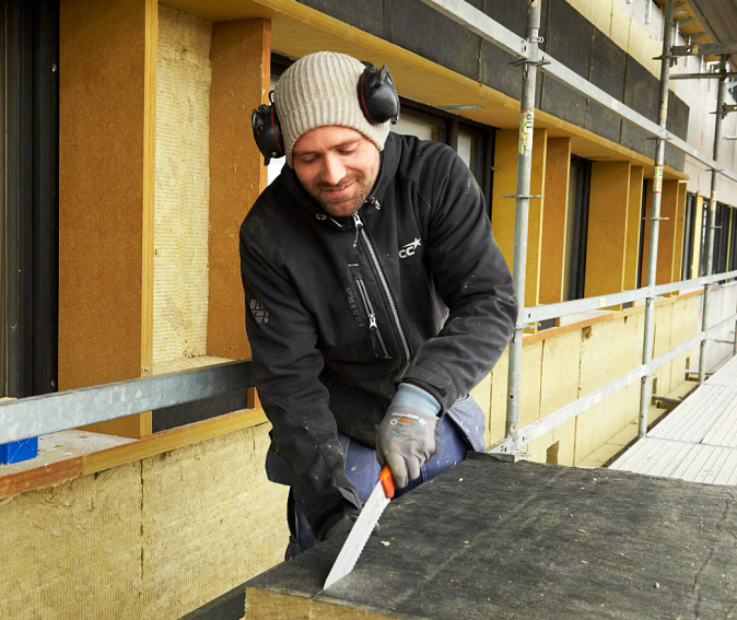 Byggarbetare i svart jacka och grå mössa med hörselskydd sågar isolerskiva vid gul fasad