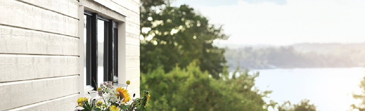 Vitt träfasadhus med fönster och färgglada blommor, grönskande träd och sjöutsikt.
