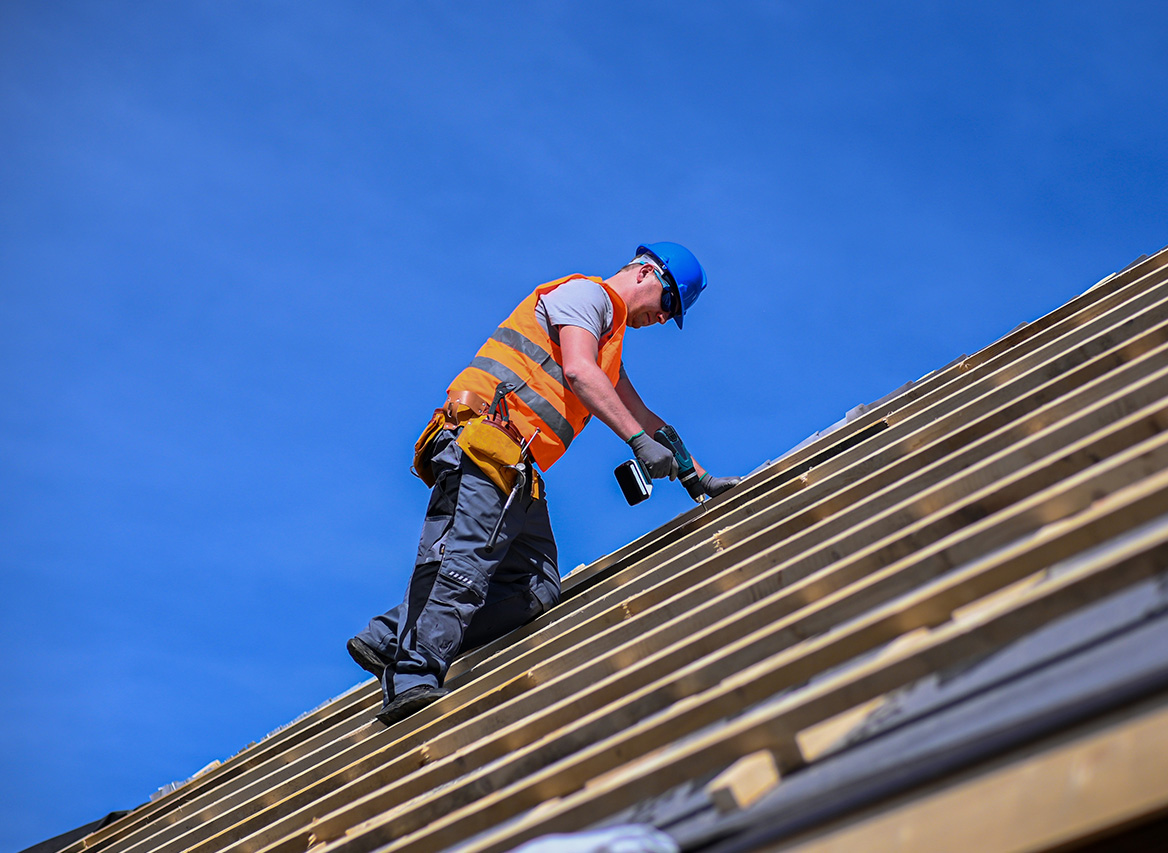 Byggarbetare i orange väst och blå hjälm borrar i trästomme på tak under klarblå himmel.