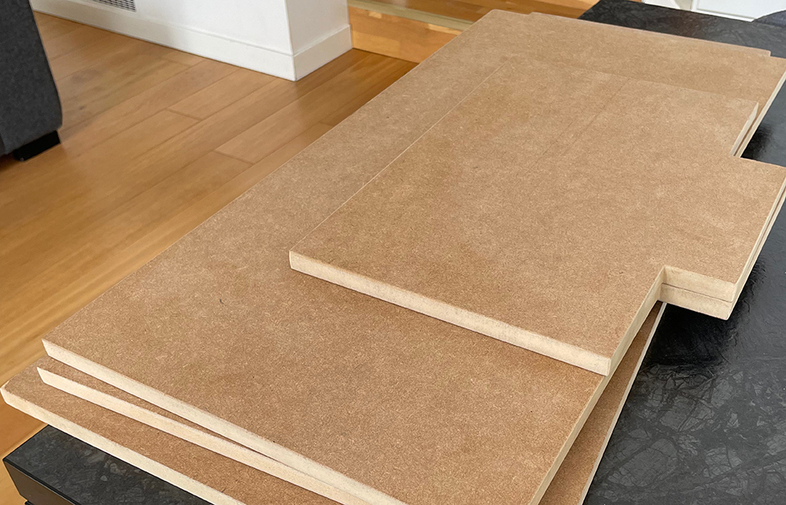 Stackade ljusbruna MDF-skivor med utskurna kanter på svart bord i rum med trägolv.