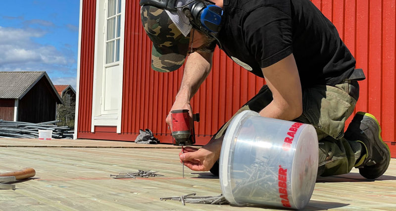 Man med kamouflagemössa och hörselskydd skruvar på ljus trätrall framför rött hus.
