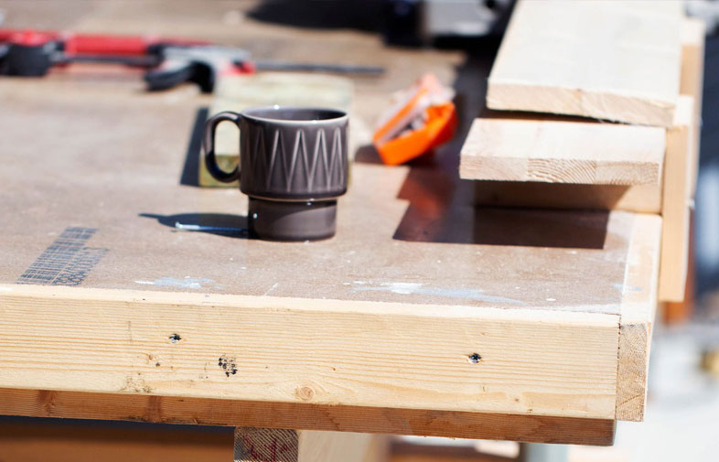 Närbild av ljus snickarbänk med mörkgrå mugg, orange kniv, verktyg och träplankor.