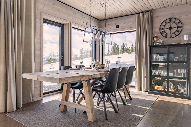 Matplats i ljus trästuga med stort bord, svarta stolar, glaslampa och snöutsikt.