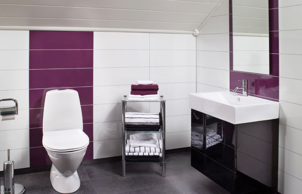 Badrum med vit toalett, handfat, lila kakelrand och hylla med vita och lila handdukar