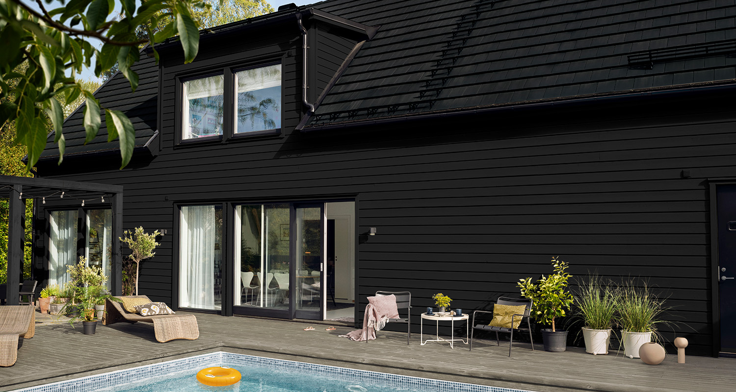 Svart trähus med stora glasdörrar, träterrass med pool, solstolar, sittgrupp och krukväxter.