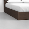 A close-up of a modern wooden bed frame with a white mattress and pillows, set on a light gray floor against a plain white wall.