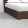 A close-up of a modern wooden bed frame with a white mattress and pillows, set on a light gray floor against a plain white wall.
