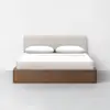 A modern bed with a light beige upholstered headboard, white pillows, white bedding, and a wooden base, set against a plain white wall and light gray floor.