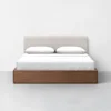 A modern bed with a light beige upholstered headboard, white pillows, white bedding, and a wooden base, set against a plain white wall and light gray floor.
