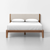 A wooden bed frame with a padded beige headboard, white bedding, and two white pillows, set against a plain white wall and white floor.
