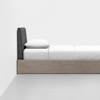 A modern, minimalist bed with a dark upholstered headboard, white bedding, and a light wood base, positioned against a plain white wall on a light gray floor.