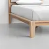 A close-up view of a wooden bed frame with a white mattress and bedding in a minimal, modern room with a light gray floor and white walls.