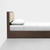 A minimalist wooden bed with a beige upholstered headboard, white pillow, and white bedding, placed against a plain white wall on a light gray floor.