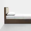 A minimalist wooden bed with a beige upholstered headboard, white pillow, and white bedding, placed against a plain white wall on a light gray floor.