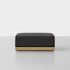 A square, dark gray ottoman with a wooden base sits on a light gray floor against a plain white background.