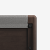 Close-up of the corner of a dark wooden crib with a soft, light gray fabric cover draped over the top rail, set against a plain white background.