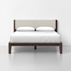 A modern bed with a dark wooden frame, light upholstered headboard, white bedding, and two white pillows, set against a plain white wall and gray floor.