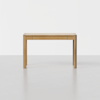 A simple rectangular wooden desk with clean lines, light brown finish, and two small drawers, standing on a light-colored floor against a plain white background.