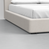 Close-up of a beige upholstered bed frame with white bedding on a light gray floor, showing the lower corner and sturdy construction.