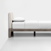 A minimalist bed with a beige upholstered headboard, white pillow, and white bedding on a wooden frame, set against a plain white wall and floor.