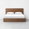 A minimalist wooden bed frame with a rectangular headboard, neatly made with white bedding and two white pillows, set against a plain white wall and gray floor.