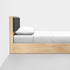 Minimalist wooden bed with a black cushioned headboard, white pillow, and neatly made white bedding, positioned against a plain white wall on a smooth gray floor.