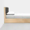 Minimalist wooden bed with a black cushioned headboard, white pillow, and neatly made white bedding, positioned against a plain white wall on a smooth gray floor.