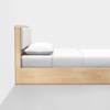 A minimalist wooden bed with a light wood frame, a padded headboard, and white bedding, including a pillow and a neatly made sheet, set against a plain white wall on a gray floor.