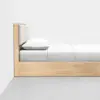 A minimalist wooden bed with a light wood frame, a padded headboard, and white bedding, including a pillow and a neatly made sheet, set against a plain white wall on a gray floor.
