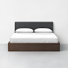 A modern bed with a dark wood frame, white bedding, two white pillows, and a dark gray upholstered headboard, positioned against a plain white wall on a light-colored floor.