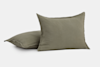 Two square olive green pillows with a simple, plain design are placed on a white background. One pillow is standing upright, while the other lies flat behind it.