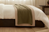 A neatly made bed with white bedding and two folded blankets, one beige and one olive green, draped across the foot. The room features light wood floors and sheer curtains letting in natural light.