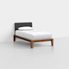A single bed with a wooden frame, white bedding, a single pillow, and a dark upholstered headboard, set against a plain white wall and floor.