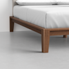 Close-up of a wooden bed frame with clean lines and tapered legs, supporting a mattress with white bedding, set on a smooth, light gray floor.