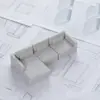 A small, gray, modular sectional sofa model sits on top of scattered technical drawings and blueprints. The model has three seats and three back cushions, fitting together on a corner.