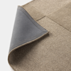 A close-up of a beige textured rug with a dark gray non-slip backing, one corner flipped over to show the underside, on a white background.