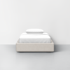 A single bed with a light beige upholstered frame, neatly made with white bedding and a single white pillow, set against a plain white wall in a minimalistic room.