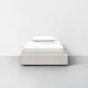 A single bed with a light beige upholstered frame, neatly made with white bedding and a single white pillow, set against a plain white wall in a minimalistic room.