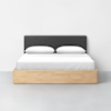 A modern bed with a light wood frame, white bedding, two pillows, and a dark gray upholstered headboard, set against a plain white wall and a light gray floor.