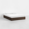 A minimalist bed with a dark wooden frame, white bedding, and two white pillows, set against a plain white wall and floor.