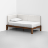 A modern daybed with a wooden frame, white mattress, and three upholstered white backrests is placed in the corner of a minimal, light gray room. A single white pillow rests on the mattress.