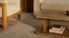A wooden coffee table with books and a candle sits on a beige carpet, next to light upholstered armchairs and a sofa in a cozy, neutral-toned living room.