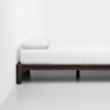 A minimalist bed with a dark wooden frame, white bedding, and a single white pillow, placed against a plain white wall on a light-colored floor.