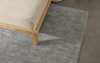 A corner of a wooden bed frame with a light-colored mattress sits on a textured gray area rug over a smooth, dark floor.