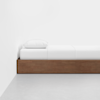 A minimalist bed with a wooden frame, white sheets, and a single white pillow positioned against a plain white wall on a light gray floor.