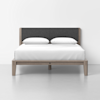 A modern bed with a wooden frame, dark gray upholstered headboard, and neatly made white bedding with two white pillows, set against a plain white wall and floor.