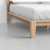 Close-up view of a wooden bed frame with a white mattress and pillows, showing the corner of the frame and leg on a smooth, light gray floor.
