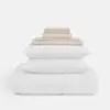 A neatly folded stack of beige bed sheets sits atop two white pillows and a white duvet, all arranged on a plain white background.