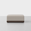 A rectangular, beige upholstered ottoman with a smooth fabric surface and a dark wooden base, placed on a light gray floor against a plain white background.