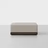 A rectangular, beige upholstered ottoman with a smooth fabric surface and a dark wooden base, placed on a light gray floor against a plain white background.